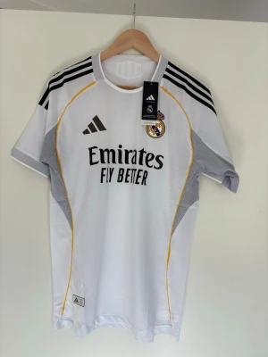 Real Madrid matchtröja Adidas M - Snygg vit Real Madrid matchtröja från Adidas med svarta detaljer och gula linjer. Tröjan har klubbmärke på bröstet, Adidas-logga och sponsortryck. Tillverkad i lätt och ventilerande funktionsmaterial med slim fit passform.