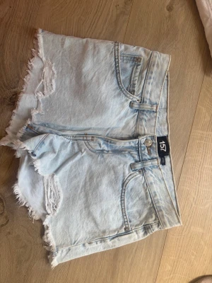 Ljusa denimshorts från 157 Xs - Säljer ett par ljusblå denimshorts från 157 med fransiga kanter. Klassisk femficksmodell med knapp och dragkedja framtill. Perfekta för sommaren och ger en avslappnad vibe.