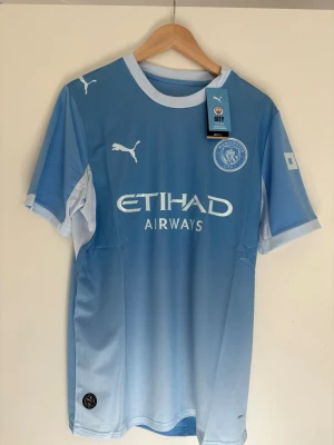 Manchester City matchtröja Puma - Ljusblå Manchester City matchtröja från Puma med vita detaljer och klubbmärke på bröstet. Tillverkad i återvunnet material (Re:Fibre) och har Etihad Airways som sponsortryck. Perfekt för fotbollsträning eller att bära till vardags.