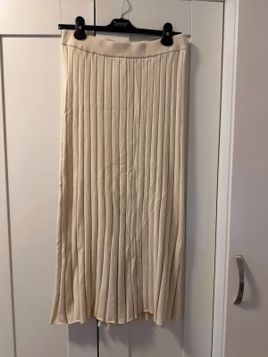 Plisserad beige långkjol - Snygg beige långkjol med plisserad design och bred resår i midjan. Kjolen har ett mjukt, stickat material som ger en skön och avslappnad känsla. Perfekt att styla med en enkel topp eller stickad tröja för en trendig look.