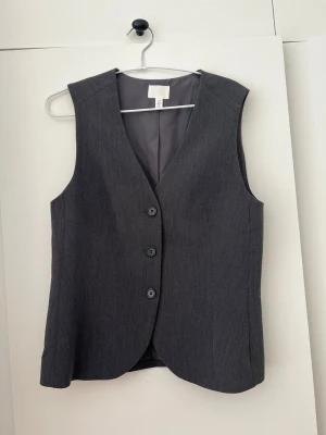 Mörkgrå kostymväst från H&M - Stilren mörkgrå kostymväst från H&M med tre knappar framtill och klassisk V-ringning. Storlek 38, använd endast en gång 🩷