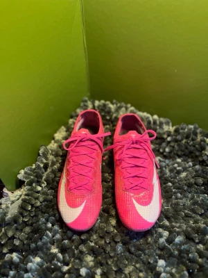 Kylian Mbappe x Mercurial Vapor 13 Elite FG 'Pink Panther' - Säljer ett par snygga fotbollsskor från Nike i en stark rosa färg med vit swoosh på ovansidan. Skorna har snörning och är tillverkade i ett lätt syntetmaterial som ger bra bollkontroll och passform. Perfekta för dig som vill sticka ut på planen.