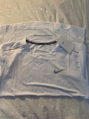 Vit Nike Running t-shirt - Säljer en vit Nike Running t-shirt med klassisk rund hals och korta ärmar. Tröjan har en diskret Nike-logga på bröstet och är tillverkad i ett lätt och ventilerande syntetmaterial som passar perfekt för löpning och träning.
