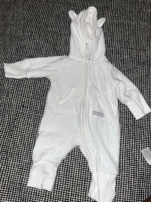 Vit overall med öron från Polarn O. Pyret - Mysig vit overall från Polarn O. Pyret med huva och söta öron. Overallen har dragkedja framtill, två fickor och är tillverkad i mjukt frottéliknande material. Perfekt för kyligare dagar och supergullig design.
