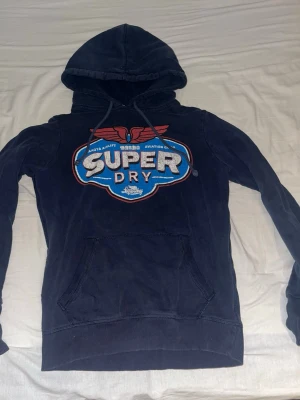 Mörkblå Superdry hoodie med tryck - Säljer en mörkblå hoodie från Superdry med stort färgglatt tryck på bröstet och broderad logga på ärmen. Hoodien har huva med snören och en magficka. Tillverkad i mjuk bomull, perfekt för en avslappnad streetwear-look.
