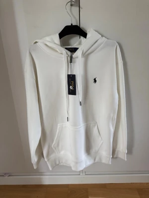 Vit hoodie från Polo Ralph Lauren - Snygg vit hoodie från Polo Ralph Lauren med klassisk huva och dragsnören. Har en stor magficka och den ikoniska broderade loggan på bröstet. Tillverkad i mjukt bomullsmaterial som känns riktigt skönt mot huden. Perfekt för en clean och stilren look.