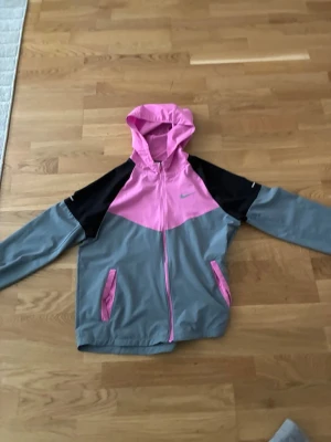 Nike vindjacka rosa/grå med huva - Snygg vindjacka från Nike i rosa, grått och svart. Jackan har huva, dragkedja framtill och två fickor med rosa detaljer. Tillverkad i lätt och vindtätt material, perfekt för blåsiga dagar. Nike-logga på bröstet för extra stilpoäng.