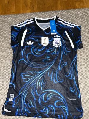 Argentina Adidas fotbollströja  - Säljer en svart och blå Argentina fotbollströja från Adidas med ClimaCool-material. Tröjan har vita detaljer, FIFA World Champions 2022-märke och AFA-logga på bröstet. Snyggt mönster i blått och vitt, perfekt för fotbollsfans.