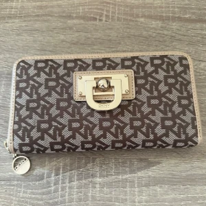 Brun DKNY plånbok med monogram - Snygg brun plånbok från DKNY med klassiskt monogrammönster i beige och silverfärgade metalldetaljer. Plånboken har dragkedja runtom och ett dekorativt spänne framtill med DKNY-logga. Tillverkad i tåligt syntetmaterial. Aldrig använd! OBS: FRAKTAR INOM 24H!🩷