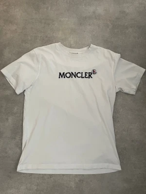 Vit Moncler t-shirt med broderad logga - Säljer en clean vit t-shirt från Moncler med stor broderad logga i svart och en liten Moncler-patch på bröstet. Klassisk rund hals och korta ärmar. Perfekt till jeans eller shorts för en enkel men snygg look.