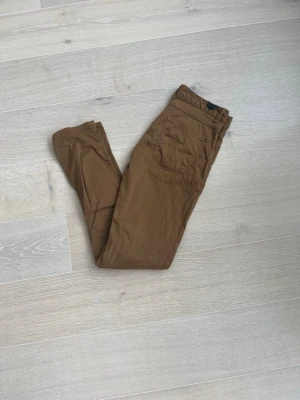 chinos - Snygga bruna chinos i slim fit-modell med klassiska fickor fram och bak. Byxorna har en enkel knappstängning och är tillverkade i ett mjukt bomullsmaterial som ger en skön känsla. Perfekta för dig som gillar en clean och stilren look.