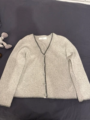 Ljusgrå kofta med svarta detaljer - Snygg ljusgrå kofta från Filippa K med svarta kantdetaljer längs v-ringningen, knäppningen och nederkanten. Koftan har lång ärm och en rak passform. Perfekt för lager-på-lager och enkel att matcha.