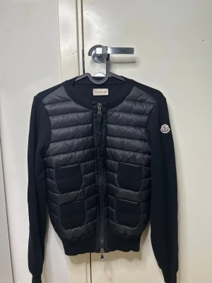 Svart cardiganjacka från Moncler - Snygg svart cardiganjacka från Moncler med quiltad front och två fickor. Jackan har rund halsringning, ribbade muddar och dragkedja framtill. Ärmarna är stickade och loggan sitter på vänster ärm. Perfekt för lager på lager och streetstyle.