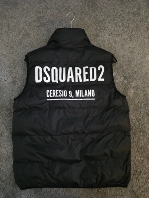 Svart dunväst från Dsquared2 - Snygg svart dunväst från Dsquared2 med hög krage och dragkedja framtill. På ryggen står det 'DSQUARED2 CERESIO 9, MILANO' i vit text. Västen har två fickor med dragkedja och quiltad design. Perfekt för lager på lager och streetwear.