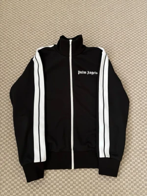 Palm Angels svart zip-tröja (M) - Svart zip-tröja från Palm Angels med vita dubbla ränder längs ärmarna och logotyp på bröstet. Hög krage och ribbade muddar. Materialet är troligtvis polyester, perfekt för en sportig och trendig look.