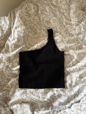 Svart oneshoulder ribbad topp - Snygg svart oneshoulder topp med ribbad struktur. Perfekt att styla till jeans eller kjol för en trendig look. Toppens asymmetriska axelband ger en cool och modern vibe.