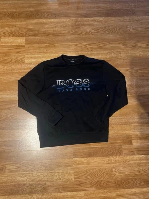 Svart sweatshirt från Hugo Boss - Svart sweatshirt från Hugo Boss med stort vitt och blått logotryck på bröstet. Tröjan har rund halsringning och ribbade muddar vid ärmslut och nederkant. Perfekt för en avslappnad och stilren look.