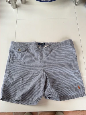 Randiga shorts från Polo Ralph Lauren - Snygga blåvit randiga shorts från Polo Ralph Lauren med klassisk passform. Shortsen har en liten broderad logga i orange vid benet och knappdetalj vid fickan. Perfekta för sommardagar och har en tidlös look.