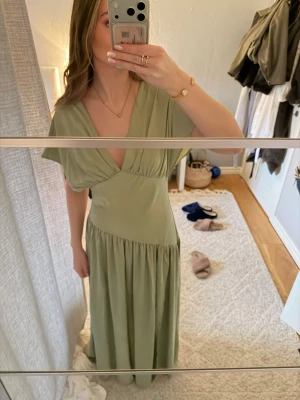 Balklänning  - Säljer en ljusgrön långklänning med omlott och djup v-ringning. Klänningen har korta ärmar och markerad midja med ett mjukt, flowy fall. Perfekt för sommarens alla tillfällen. Materialet känns lätt och svalt.
