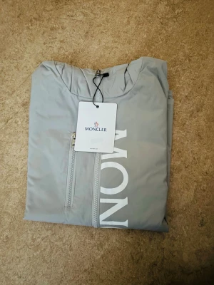 Moncler Windbreaker - Snygg ljusgrå vindjacka från Moncler med stor vit logga framtill. Jackan har dragkedja, två bröstfickor med dragkedja och huva. Tillverkad i ett lätt och vindtätt material som passar perfekt för blåsiga dagar. Minimalistisk design med exklusiv känsla.