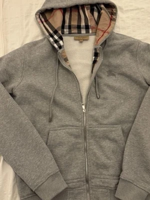 Grå zip hoodie från Burberry - Säljer en grå hoodie från Burberry med dragkedja och klassiskt rutigt foder i huvan. Hoodien har dragsko i huvan, två fickor framtill och ribbade muddar. Perfekt för dig som gillar stilrena plagg med en twist.