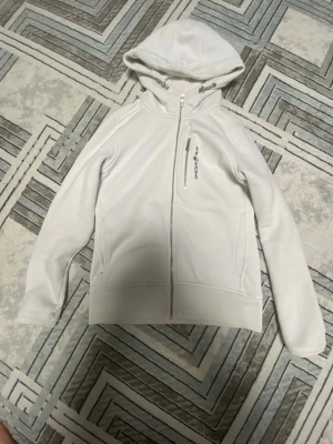 Vit hoodie från SOC med dragkedja - Snygg vit hoodie från SOC med hel dragkedja och huva. Tröjan har en bröstficka med dragkedja och diskret logga på bröstet. Perfekt för en sportig och clean look. Tillverkad i mjukt och stretchigt material för extra komfort.