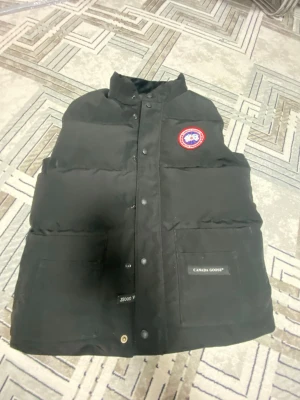 Svart dunväst från Canada Goose - Snygg svart dunväst från Canada Goose med klassisk logga på bröstet och två stora fickor framtill. Västen har hög krage, tryckknappar och dragkedja. Perfekt för lager-på-lager och håller dig varm med sitt fluffiga dunfoder.