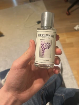 Sørensen 26.0 parfymflaska - Unik parfymflaska från Sørensen med modern design. Flaskan är genomskinlig med en silvrig metallkork och har en etikett med lila grafiskt mönster och svart text. Perfekt för dig som gillar stilrena och kreativa dofter. Tagit kanske slut på 2ml knappt använt 
