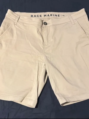 Beige shorts från Race Marine - Fin beige shorts från Race Marine med klassisk passform. De har två sidofickor, bälteshällor och en bakficka med Race Marine-logga på. Perfekta för varma dagar och enkla att matcha med. 
