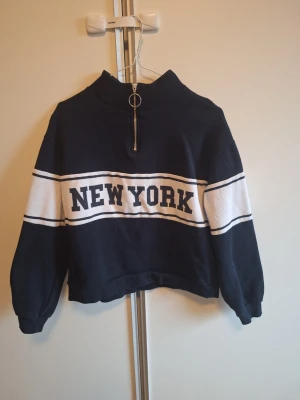 Mörkblå New York hoodie från H&M strl S - Snygg mörkblå hoodie från H&M med texten NEW YORK över bröstet. Tröjan har halv dragkedja med ringdetalj och liten krage
