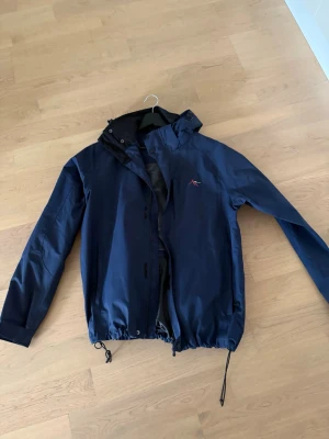 Blå vindjacka från High Mountain - Snygg marinblå vindjacka från High Mountain med huva och dragsko i nederkant. Jackan har diskret logga på bröstet och meshfoder på insidan för extra komfort. Perfekt för blåsiga dagar och enkel att matcha med olika outfits.