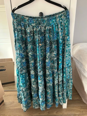 Blommig blå långkjol med snörning - Säljer en långkjol med blommigt och paisleyinspirerat mönster i blått, turkost och vitt. Kjolen har resår i midjan och snörning med träpärlor för extra detalj. Från design by si i en av sommarens trendigaste färger🦋Passar både S och M. Perfekt för sommar och utomlands!