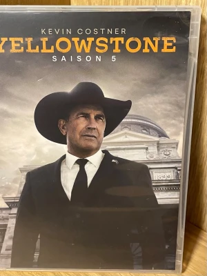 Yellowstone Säsong 5 DVD-box - Säljer en DVD-box med säsong 5 av Yellowstone. Omslaget har en cowboy i svart kostym och hatt framför en klassisk byggnad. Boxen innehåller 7 DVD-skivor och över 3 timmar extramaterial. Text på franska och engelska. Perfekt för dig som gillar drama och western.