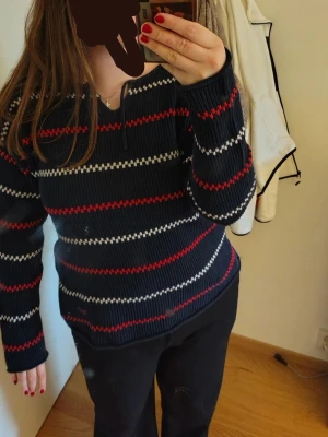 Mörkblå stickad tröja med ränder/pippitröja - Mysig mörkblå stickad tröja med röda och vita horisontella ränder. Tröjan har långa ärmar och snörning vid halsringningen för en cool detalj. Perfekt för dig som gillar klassiska färgkombinationer och vill ha en skön vardagströja.