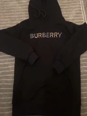 Svart hoodie - Snygg svart hoodie från Burberry med stor logga i vitt och beige på bröstet. Klassisk huva med dragsko och känguruficka framtill. Tillverkad i mjukt material som känns skönt mot huden. Perfekt för dig som vill ha en stilren och exklusiv look.