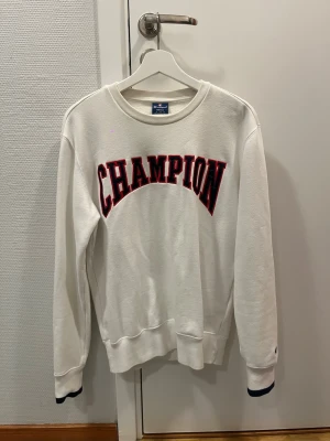 Vit sweatshirt från Champion - Vit sweatshirt från Champion med stor CHAMPION-text i rött och svart broderi över bröstet. Klassisk rund halsringning och ribbade muddar vid ärmar och nederkant. Perfekt för en avslappnad och sportig stil.