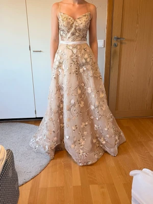 Champagnefärgad långklänning med spets - Elegant långklänning i champagnefärg med tunna axelband och v-ringning. Klänningen har ett vackert blommigt spetsmönster och markerad midja med ett satinband. Perfekt för bal eller student. Materialet känns lyxigt och skimrande. Axelbanden går att ta bort. Klänningen är både insydd och uppsydd till en 32 men går att sprätta upp till en 36.