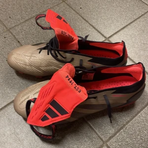 Adidas Predator fotbollsskor - Säljer ett par Adidas Predator fotbollsskor i beige och röd färg. Skorna har en modern design med snörning och är perfekta för fotbollsplanen. De har en slitstark sula med dobbar för optimalt grepp.