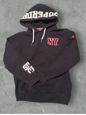 Svart hoodie med NY-tryck - Snygg svart hoodie från Superdry med vit och röd NY-brodyr på bröstet och stort Superdry-tryck på huvan. Hoodien har dragsko i huvan och en klassisk känguruficka framtill. Perfekt för en avslappnad streetwear-look.