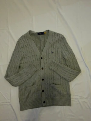 Grå kabelstickad kofta Polo Ralph Lauren - Grå kabelstickad cardigan från Polo Ralph Lauren. Tröjan är i mycket gott skick utan synliga defekter, fläckar eller slitage. Detaljer: 	•	Märke: Polo Ralph Lauren 	•	Modell: Cable Knit Cardigan 	•	Färg: Grå melange med marinblå inbrodererad pony-logga 	•	Storlek: S 	•	Passform: Regular 	•	V-ringning med bruna hornknappar 	•	Två fickor 	•	Material: Bomull Skick: Mycket gott — inga defekter.                     Modellen: 178 cm 