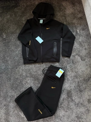 Svart Nike NOCTA hoodie med dragkedja - Svart hoodie från Nike NOCTA med dragkedja och huva. Gult Nike Swoosh på bröstet och NOCTA-tryck på ärmen. Två fickor med dragkedja framtill. Materialet är mjukt och stretchigt, perfekt för en avslappnad och cool look.