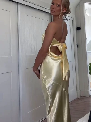 Elegant gul satinklänning från Lucy in the Sky - Gul satinklänning från Lucy in the Sky Elegant, axelbandslös maxiklänning i glansigt satin med figurnära passform och knytning i rygg. Perfekt till fest eller bal Helt ny – aldrig använd, fortfarande i originalförpackning. 