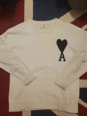 Vit stickad tröja från Ami Paris - Vit stickad tröja från Ami Paris med svart hjärta och bokstaven A broderat på bröstet. Tröjan har rund halsringning, långa ärmar och ribbade muddar vid ärmslut och nederkant. Perfekt för dig som gillar stilrena och trendiga plagg.