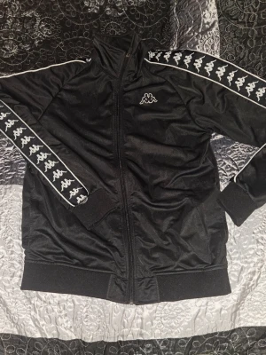 Svart Kappa ziptröja med logga - Säljer en svart ziptröja från Kappa med vit logga på bröstet och klassiska Kappa-band längs ärmarna. Tröjan har hög krage, muddar vid ärmslut och nederkant samt hel dragkedja framtill. Perfekt för en sportig och avslappnad stil.