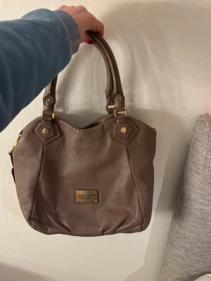 Brun handväska från Marc by Marc Jacobs - Snygg brun handväska i mjukt läder från Marc by Marc Jacobs. Väskan har dubbla handtag, avtagbar axelrem och guldfärgade metalldetaljer. Klassisk och rymlig modell med logoplatta framtill. Perfekt för dig som vill ha både stil och funktion. Pris kan diskuteras