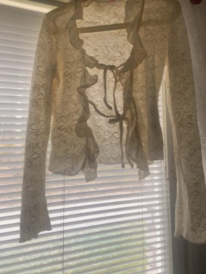 Beige spetsblus med volang och knyt - Säljer en beige, långärmad spetsblus med volangkant längs öppningen och knyt framtill. Blusen har ett blommigt spetsmönster och är lätt transparent, perfekt för en trendig och romantisk look.