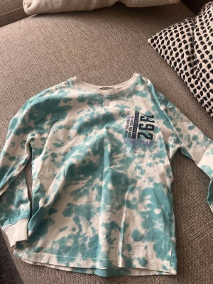 Turkos tie-dye tröja från H&M - Cool långärmad tröja från H&M i turkos och vit tie-dye. Tryck med siffran 292 och text på bröstet. Tillverkad i mjuk bomull med ribbad halsringning och muddar vid ärmslut. Perfekt för en avslappnad streetstyle-look.