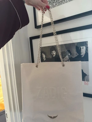 Beige shoppingväska från Zadig & Voltaire - Stilren shoppingväska i beige canvas från Zadig & Voltaire med präglad logga framtill och en liten metallvinge. Väskan har rejäla kedjehandtag i silverfärg som ger en cool touch. Perfekt för dig som vill ha en rymlig och trendig tote till vardagen.