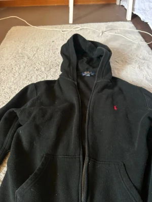 Svart hoodie från Polo Ralph Lauren - Klassisk svart hoodie från Polo Ralph Lauren med dragkedja framtill och huva. Tröjan har två fickor och den ikoniska röda logotypen broderad på bröstet. Tillverkad i mjukt bomullsmaterial som känns skönt mot huden. Perfekt för en avslappnad och stilren look.