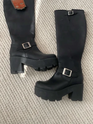 Svarta chunky platåstövlar med spänne - Säljer ett par nya , oanvända  svarta höga stövlar med chunky platå och grov sula. Stövlarna har två silvriga spännen, ett vid ankeln och ett upptill, samt dragkedja baktill. Tillverkade i ett matt, slätt material som ger en cool och edgy look. Stl 38 . 